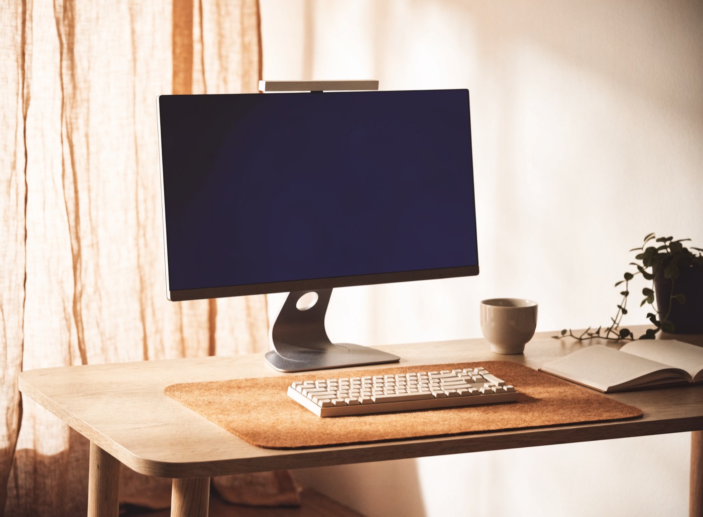 A warm minimal home office desk: monitor with a light bar, cream keyboard on a cork mat, mug, open notebook and a trailing plant