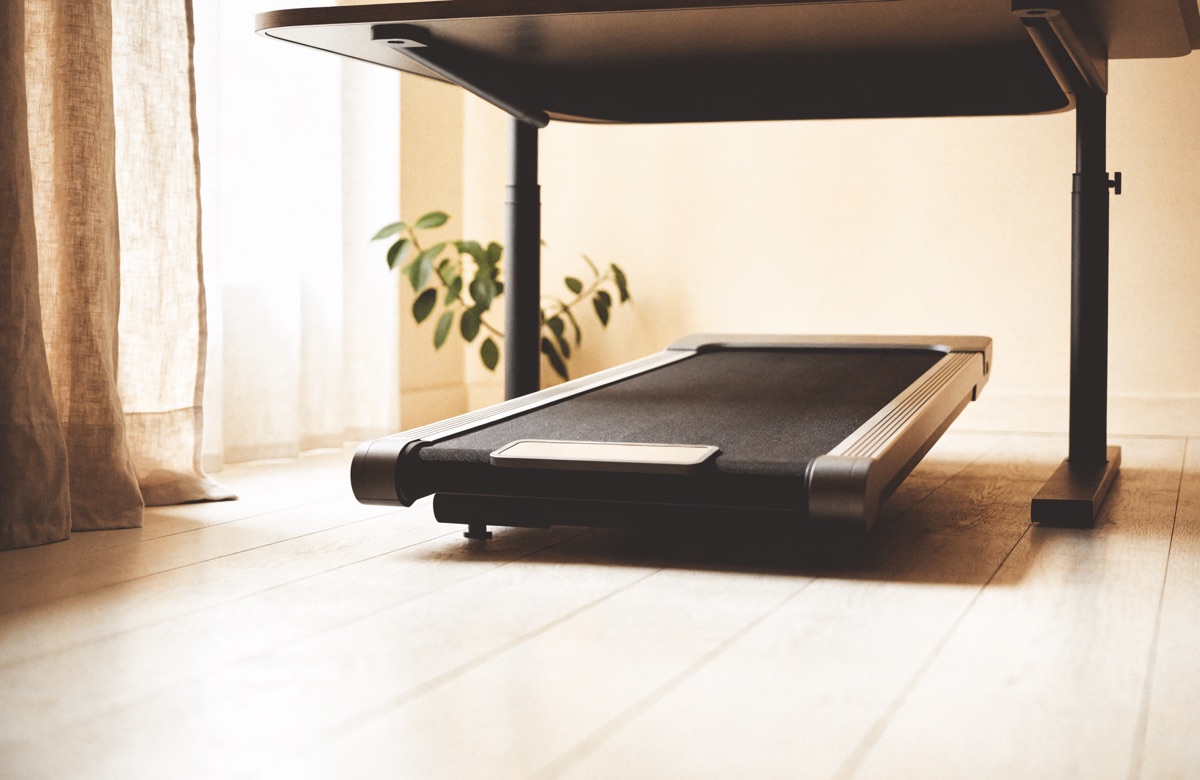 A walking pad tucked beneath a raised standing desk in a sunlit room