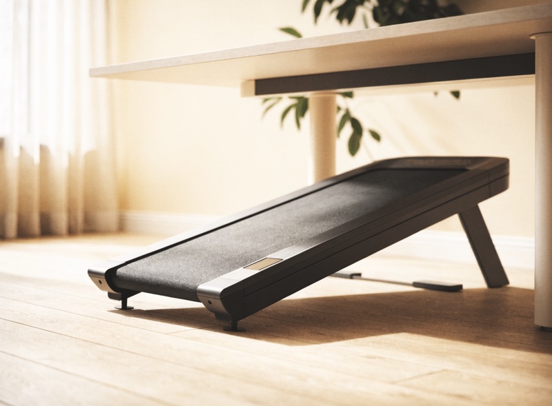 A slim walking pad on a wooden floor beneath a standing desk